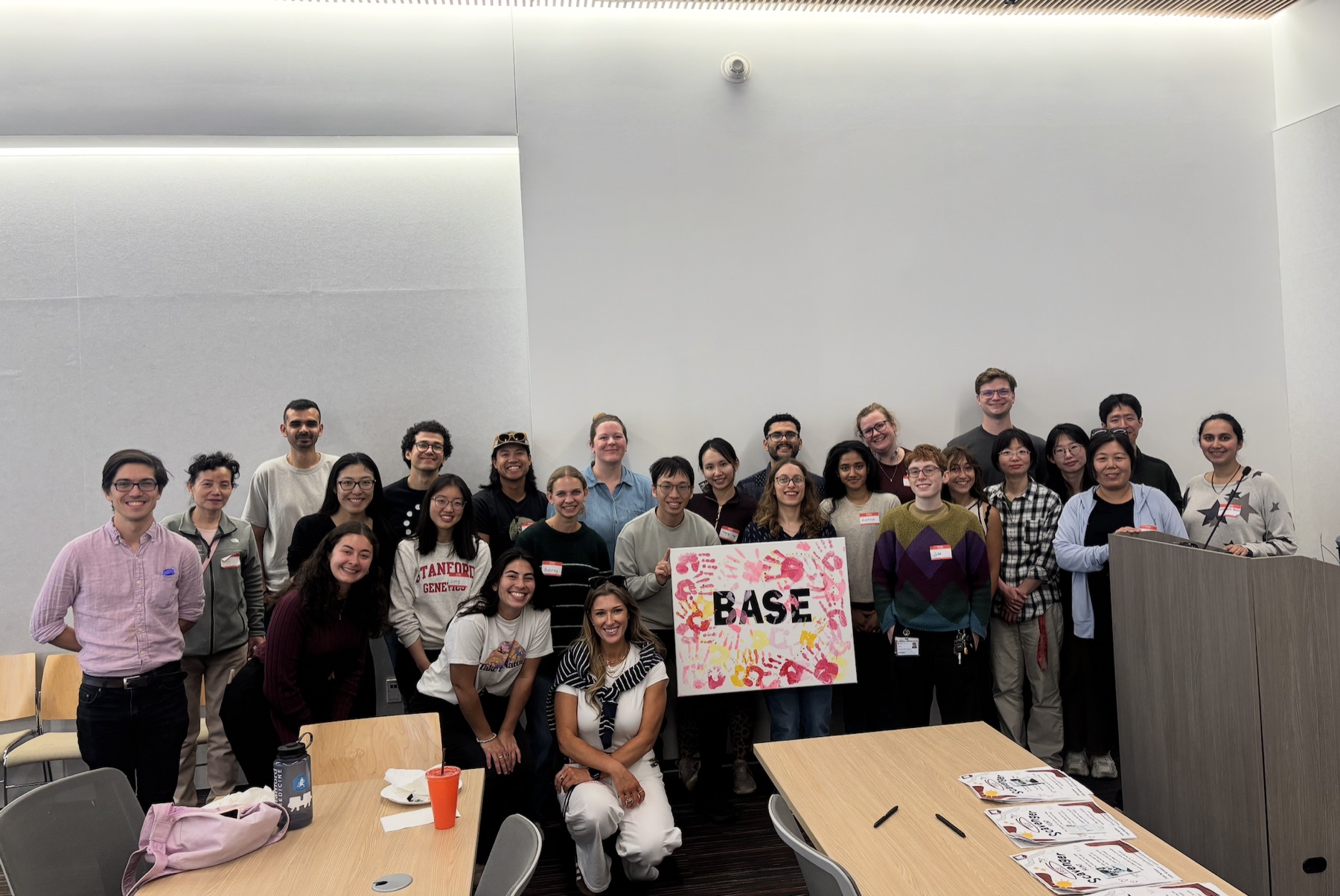 A large group of smiling people in a seminar room, two of them holding a white canvas covered in red, pink and yellow handprints with the word BASE in the middle.