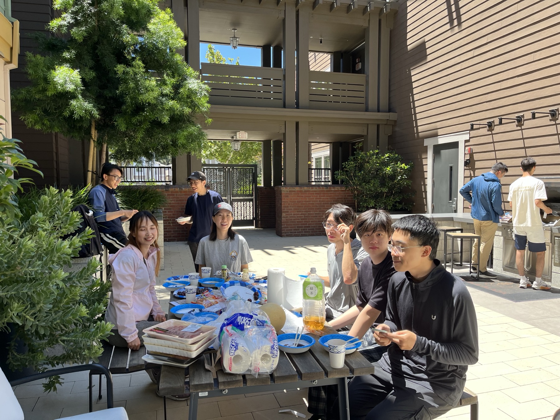 Seven people seated around a picnic table full of food in a sunny courtyard, with two more cooking at a grill in the background.