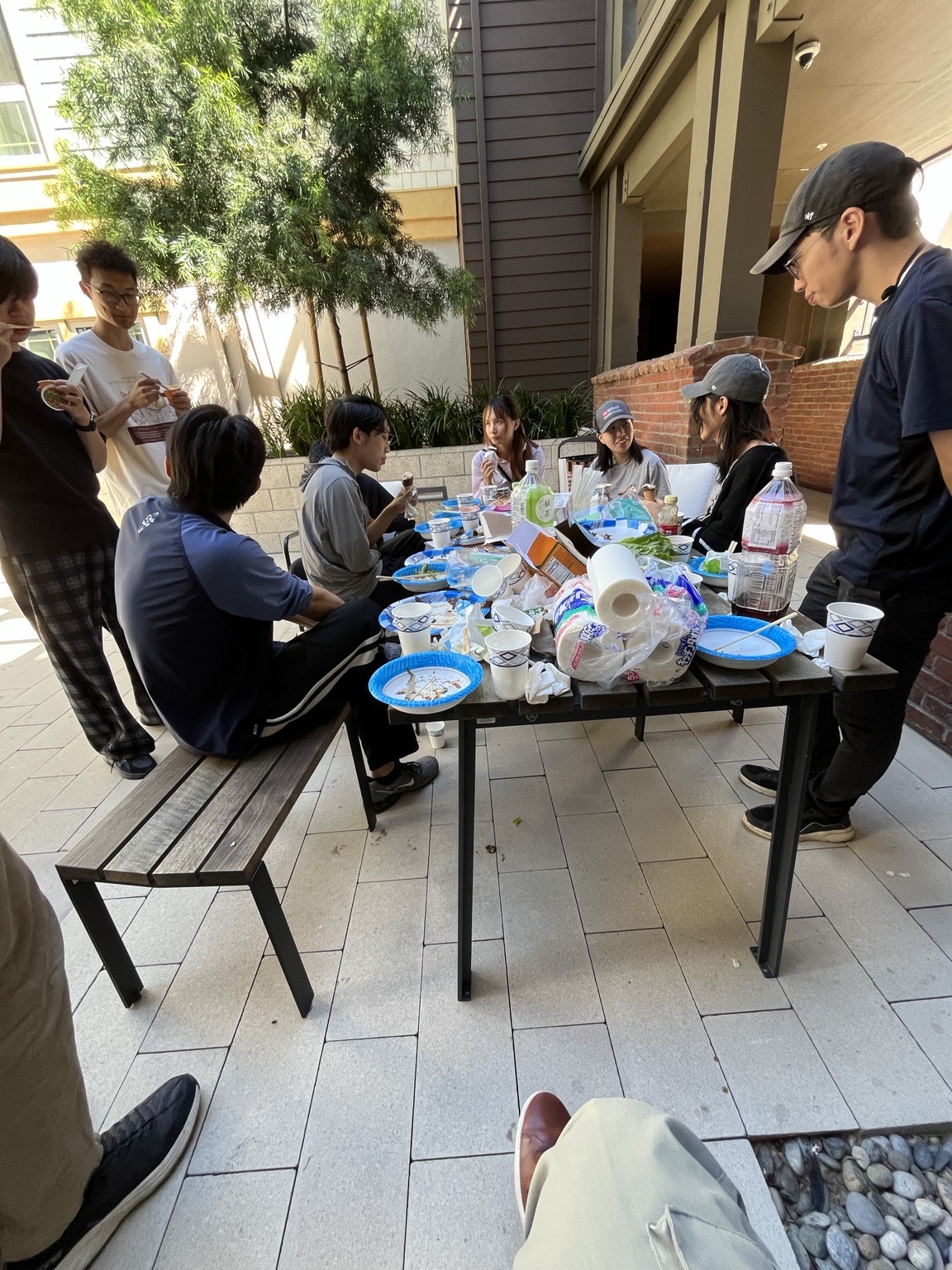 Lab members standing and sitting around a picnic table covered in plates and bottles after the meal, in a paved courtyard.