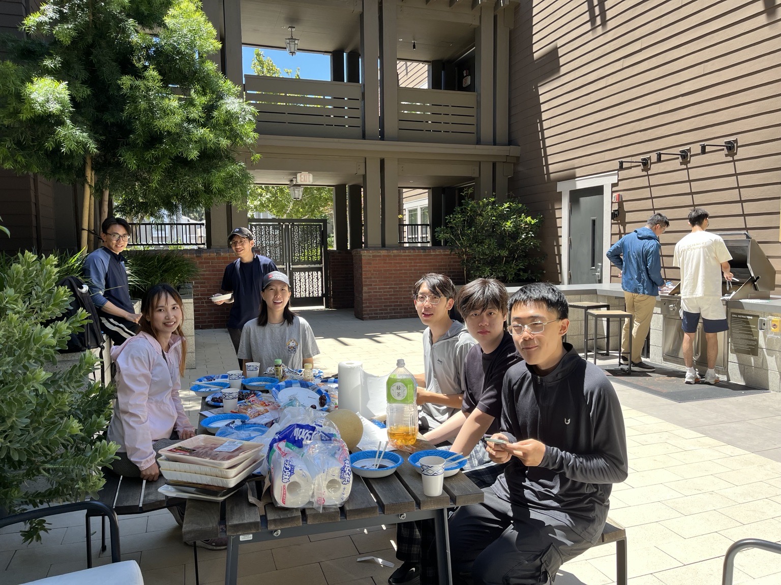 A closer view of the same picnic table, with trays of food, blue paper plates and a melon, and lab members smiling at the camera.