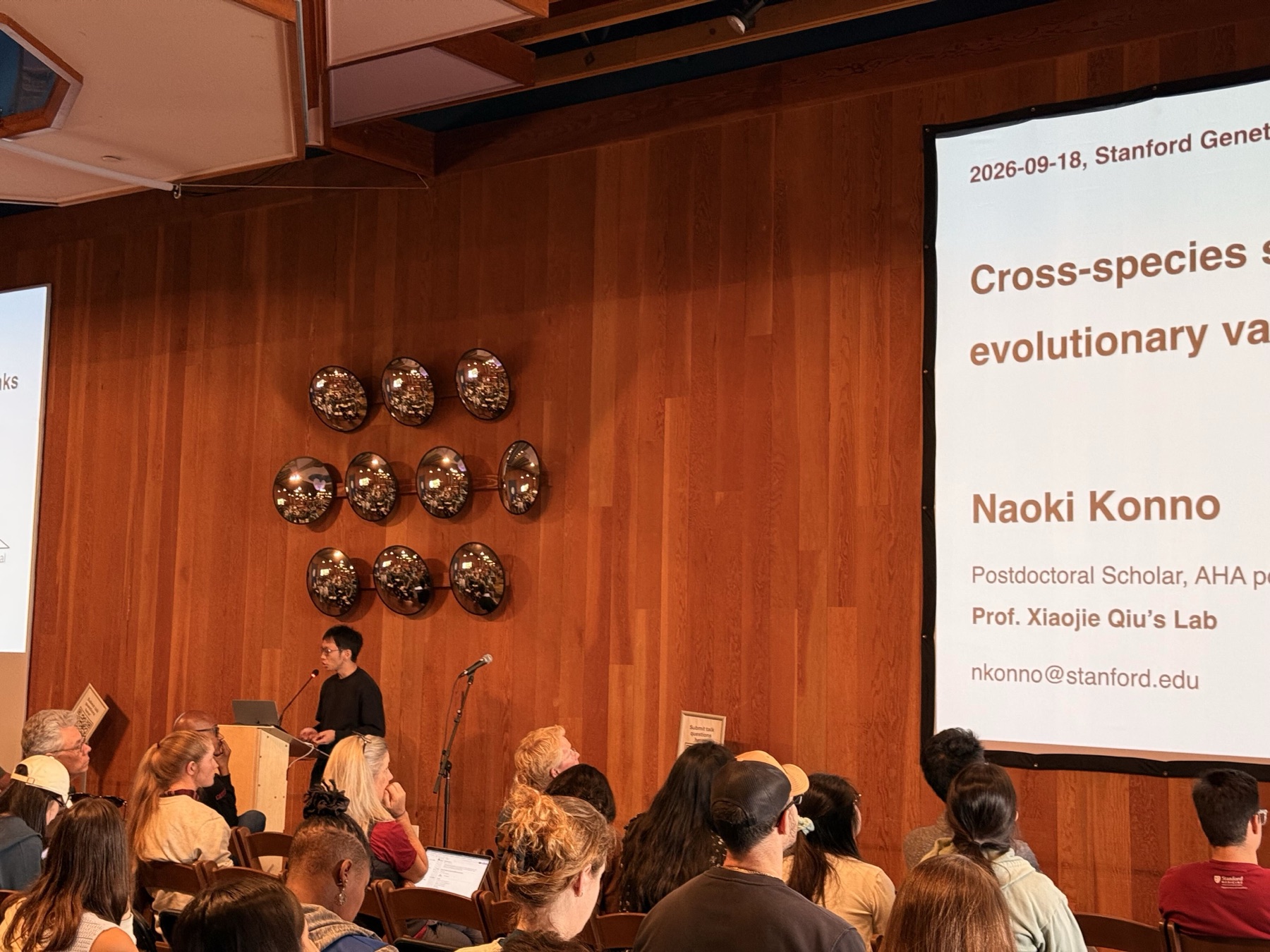 Naoki Konno speaking at a podium in a wood-panelled hall, with a projected title slide to the right and an audience seated in the foreground.