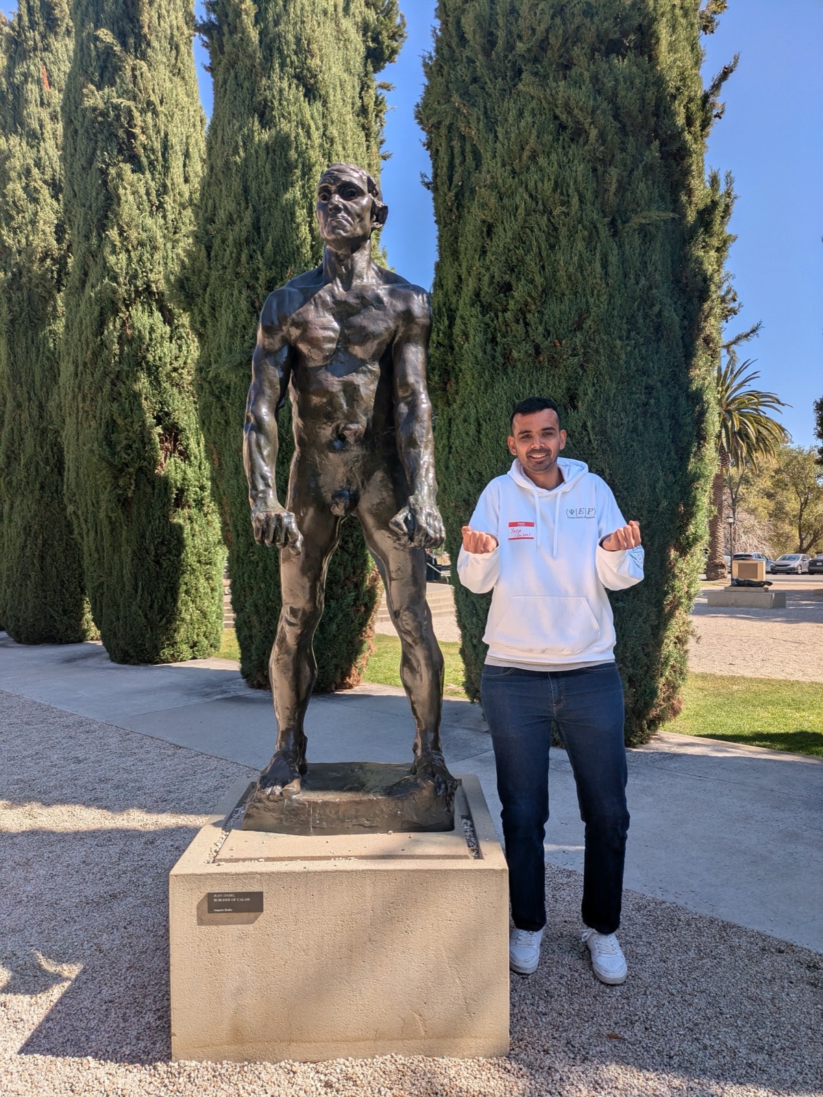 Yash Rana standing beside the bronze statue of Jean d'Aire, clenching both fists to mirror the statue's pose, in front of tall cypress trees.