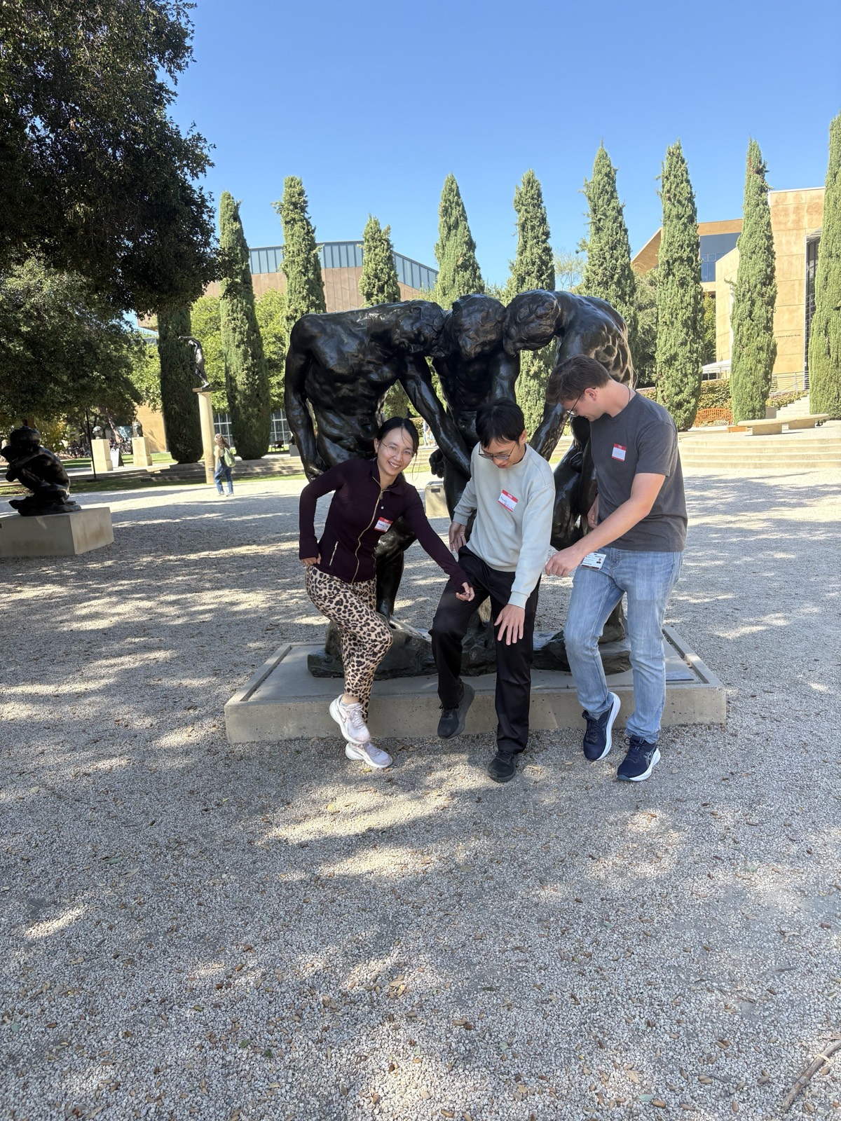 Three lab members posing in front of Rodin's bronze sculpture The Three Shades, copying its hunched, arm-linked pose, with cypress trees behind them.