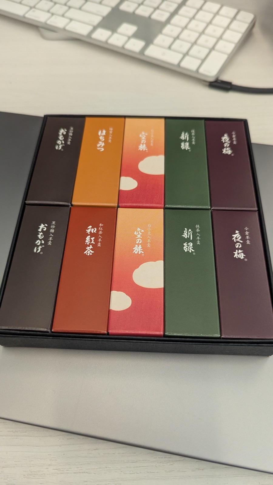 An open gift box of ten colourful yōkan bars with Japanese calligraphy labels, sitting on a desk in front of a keyboard.